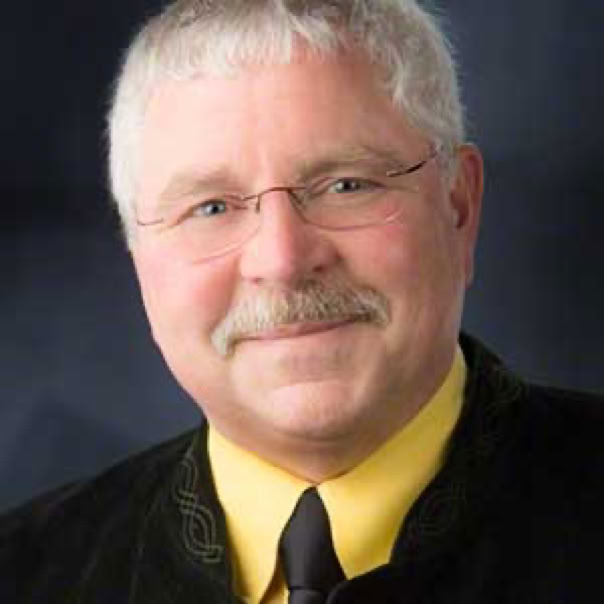 A man with gray hair and glasses, wearing a yellow shirt and a dark jacket, smiling against a dark background.